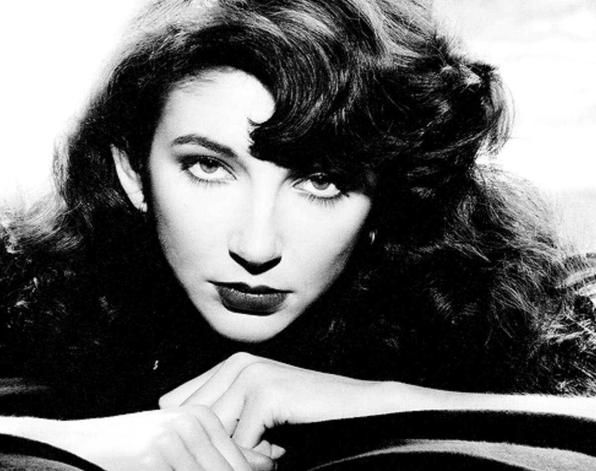 Black and white portrait of a woman with wavy dark hair, dark lipstick, and bold eyebrows, resting her chin on her hands and looking directly at the camera with an intense expression.