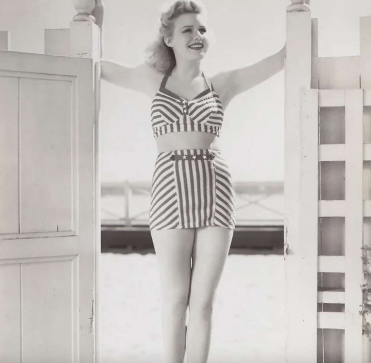 A woman in a vintage striped bikini smiles while standing between an open gate on a beach, with sand and a fence in the background. The image is in black and white.