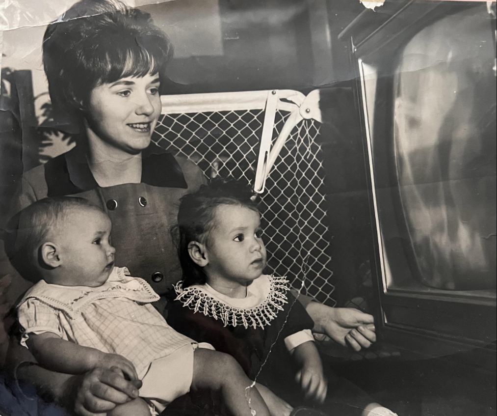 A woman sits with two young children, one in her arms and the other by her side, all watching an old-fashioned television. The photo is in black and white, giving it a vintage feel.