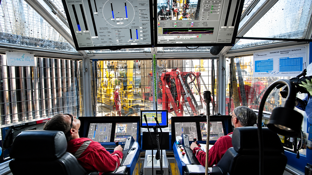 Oil Rig. Two operators in red uniforms control drilling machinery from inside a high-tech cabin with multiple screens, while workers and equipment are visible through large windows outside.