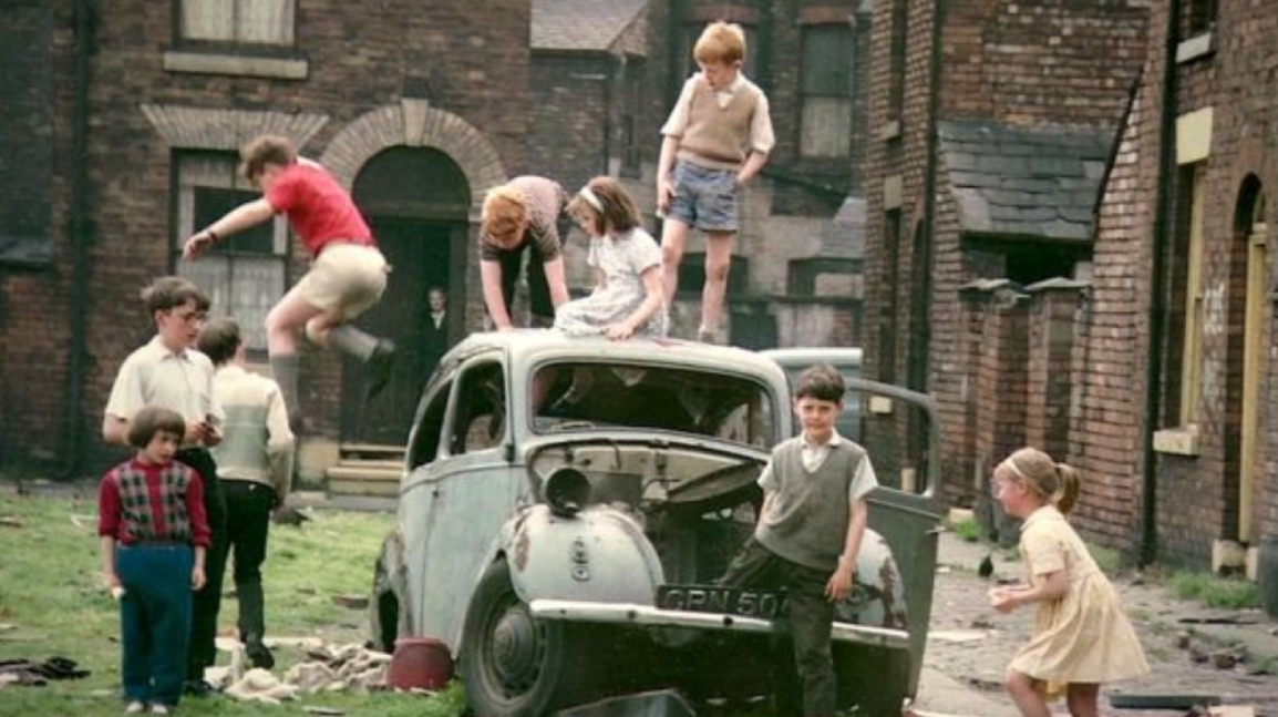 A group of children in casual clothes play on and around an old, damaged car in a grassy area between brick buildings, with some kids climbing and others standing or watching.