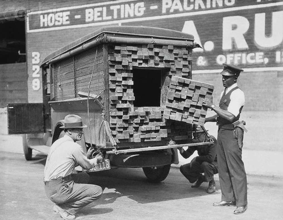 Three men inspect a truck loaded with stacked wooden planks that conceal a hidden compartment; one man removes planks to reveal the secret space as others look on and take notes.