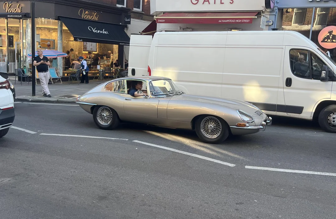 A classic silver Jaguar E-Type car drives down a city street, passing a white van. People sit outside a café and ice cream shop in the background, enjoying the sunny day.