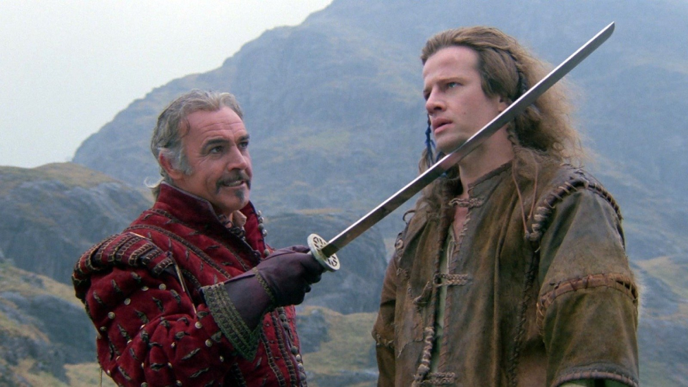 A man in ornate red clothing holds a sword to the neck of another man wearing a brown tunic, with rocky hills in the background, both looking serious.
