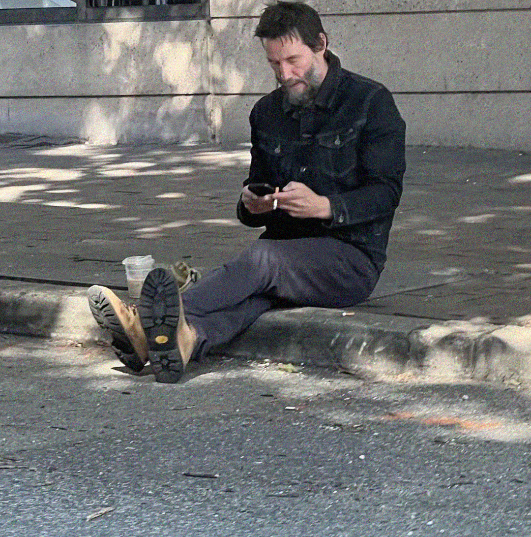 A man with a beard sits on a curb, legs stretched out, looking at his phone. He wears a dark jacket, brown boots, and has a coffee cup next to him on the sidewalk.