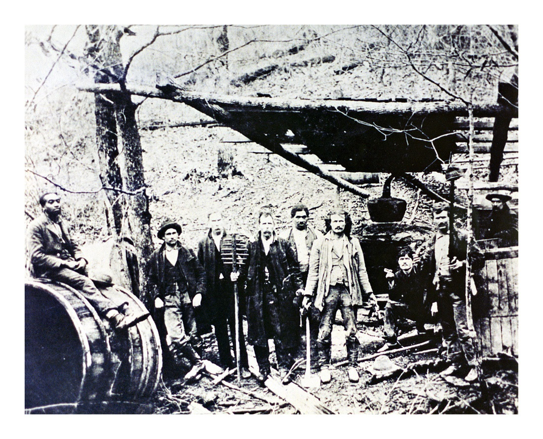 A group of men stand in a wooded area next to a large barrel and rustic wooden structures, possibly involved in early 20th-century moonshining or distilling activities. The scene appears old and weathered.