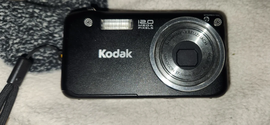 A black Kodak digital camera with "12.0 Mega Pixels" label is placed on a white surface, showing its lens, flash, and wrist strap. A textured grey fabric is partially visible in the background.