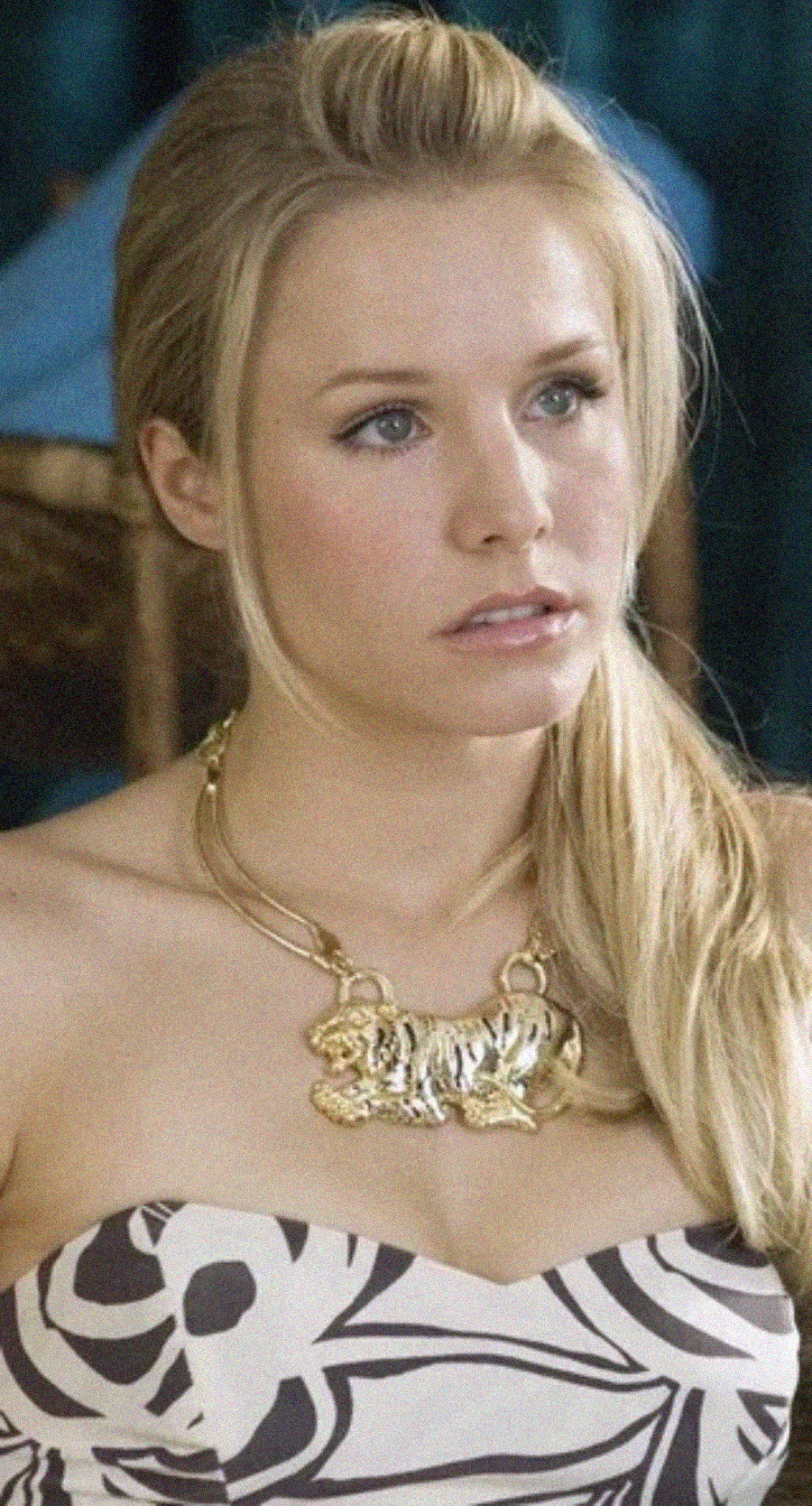 A woman with long blonde hair in a ponytail wears a strapless dress with a black and white leaf pattern and a large gold animal-shaped necklace. She has a serious expression and sits indoors.