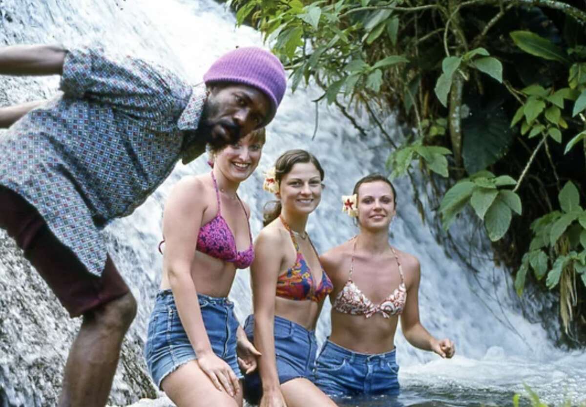 Four people pose by a lush waterfall; three women in bikinis and denim shorts sit in the water, each with a flower behind their ear, while a smiling man in a purple beanie and patterned shirt leans into the frame.