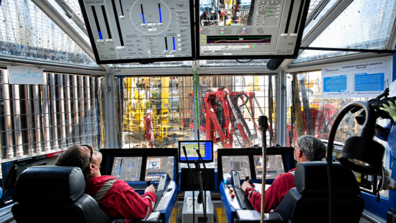 Two workers in red uniforms operate control panels inside a glass-enclosed room with multiple screens, overlooking industrial machinery and equipment outside.