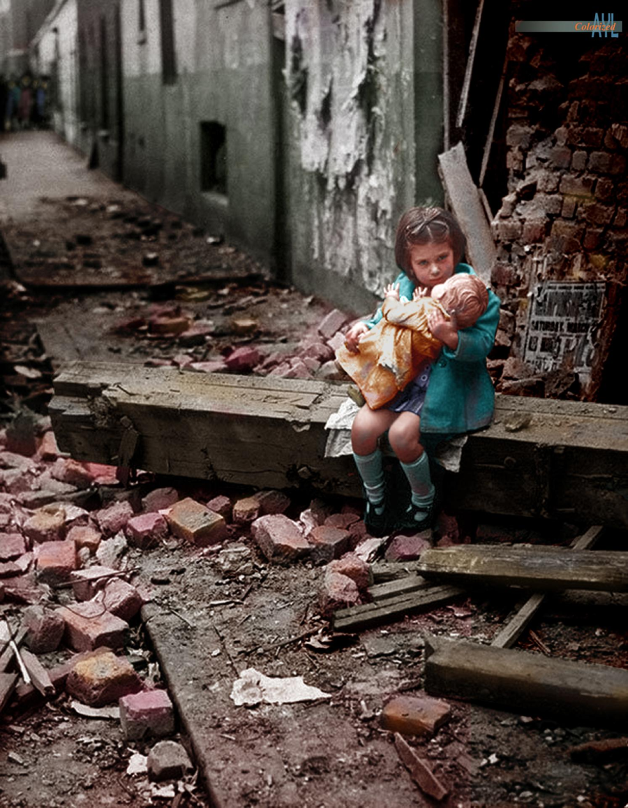 A young girl in a blue coat sits on rubble in a destroyed alley, clutching a doll. Broken bricks and debris surround her, and damaged buildings are visible in the background.