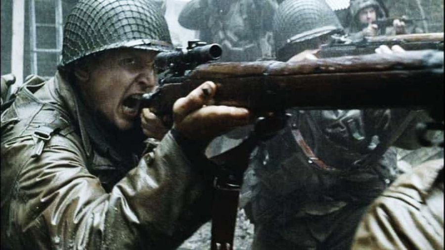 A soldier in a helmet and uniform shouts while aiming a rifle, surrounded by other soldiers in intense combat during a battle in rainy, gritty conditions.