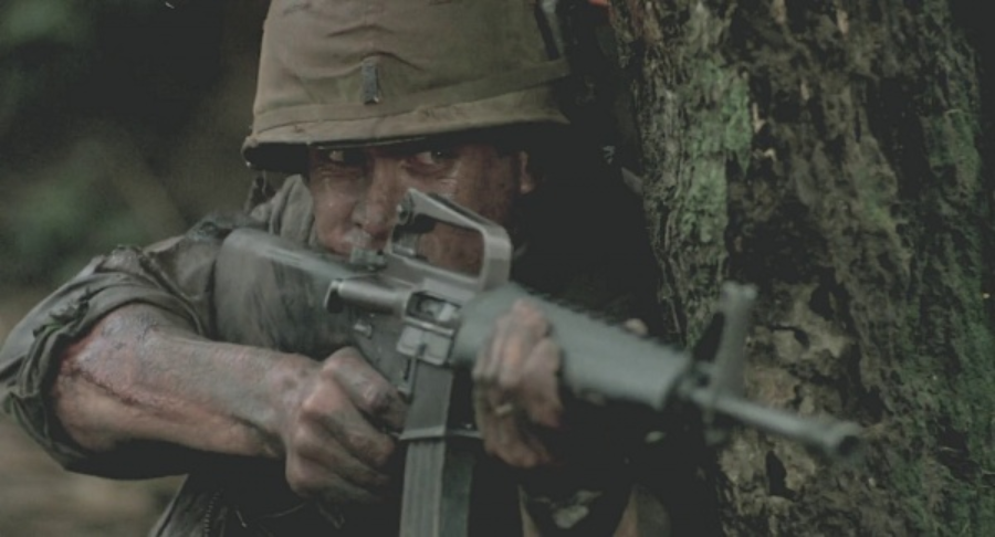 A soldier in a helmet and dirty uniform crouches behind a mossy tree, aiming a rifle with intense focus. The scene is gritty, with visible mud and wounds on his arms, creating a tense, combat atmosphere.
