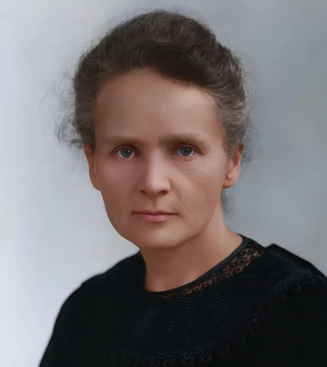A woman with light skin, blue eyes, and grayish-brown hair worn up, dressed in a black high-necked top with delicate detailing, poses against a pale, blurred background.
