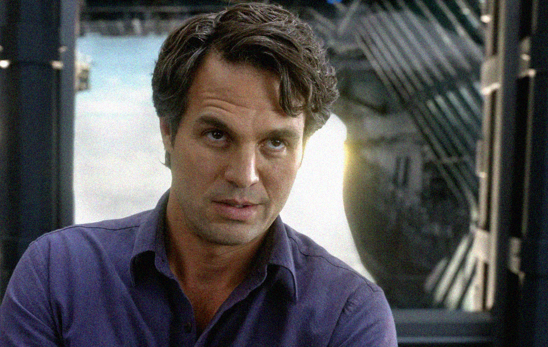 A man with wavy brown hair in a purple shirt looks to the side with a serious expression. He is indoors with futuristic metallic structures and large windows in the background.