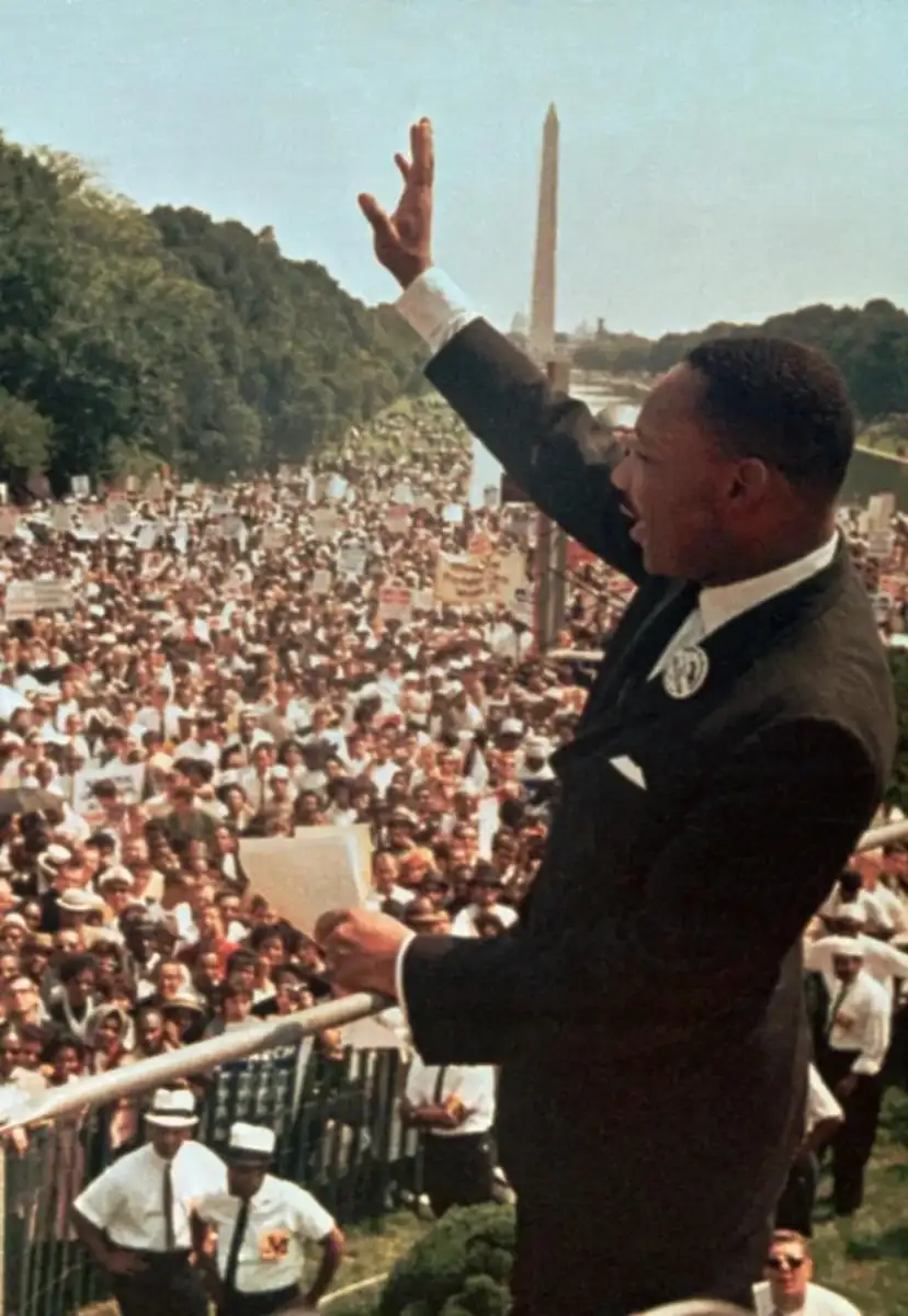 A man in a suit stands on a platform, raising his hand and holding papers, addressing a large crowd gathered outside near the Washington Monument.