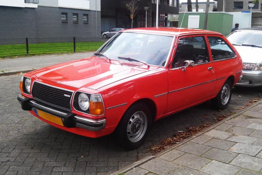 A bright red vintage hatchback car is parked on a city street beside a sidewalk, with buildings and another parked car visible in the background.