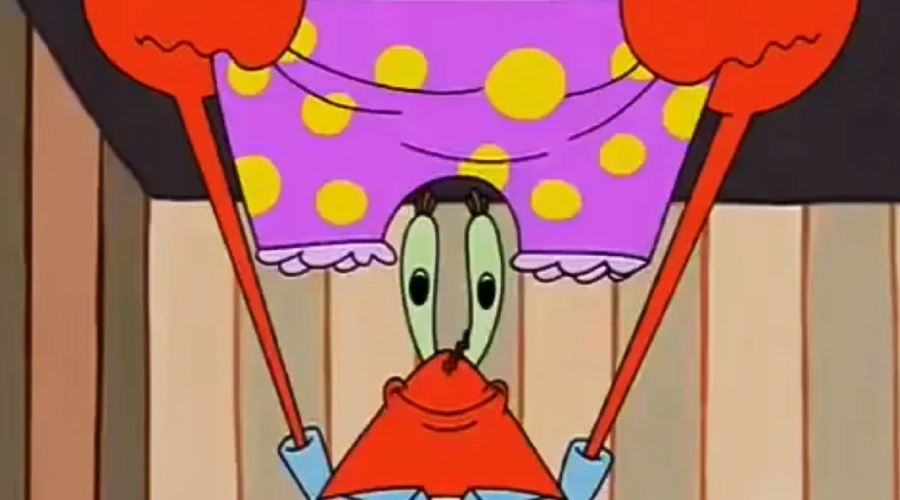 A cartoon crab character holds up a pair of large, purple boxer shorts with yellow polka dots, smiling while standing indoors against a wooden wall.