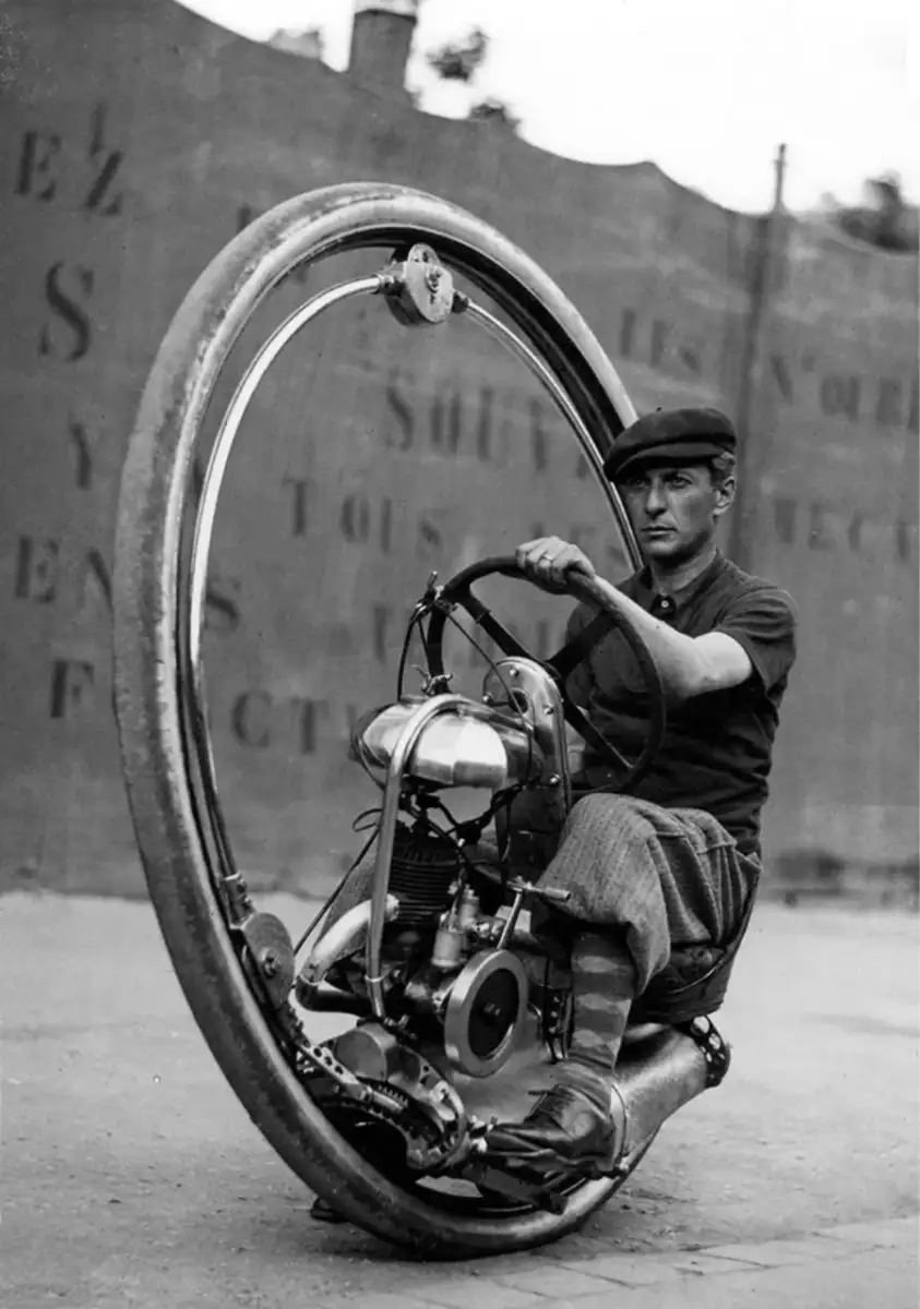 A man wearing a cap and knickerbockers rides a vintage, one-wheeled monowheel motorcycle on a paved street, with a wall bearing faint, partially visible text in the background.