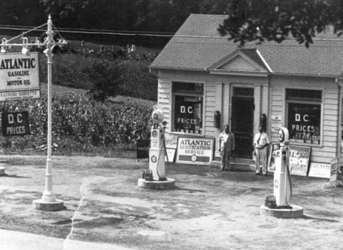 Black-and-white photo of a vintage gas station with two old-style pumps, an "Atlantic Gasoline" sign, and a small building. Two people stand near the entrance. Signs advertise gas prices and lubrication service.