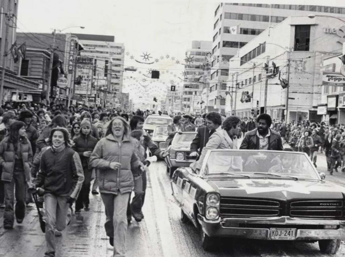 A black-and-white photo showing a large crowd celebrating on a city street. People run alongside a convertible car, with high-rise buildings in the background and festive decorations overhead.