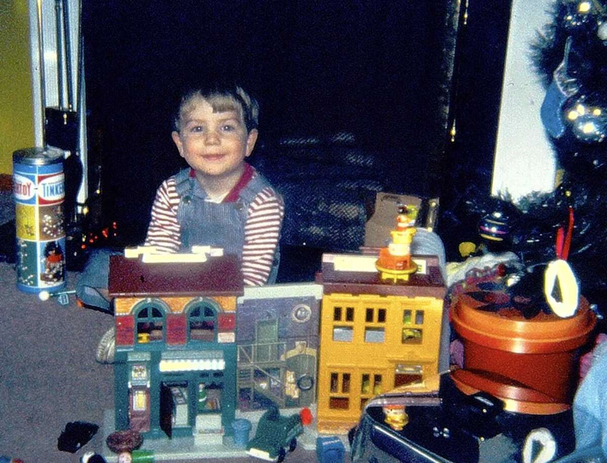 A young child in striped shirt and overalls smiles while sitting on the floor surrounded by toy buildings and holiday decorations next to a Christmas tree.