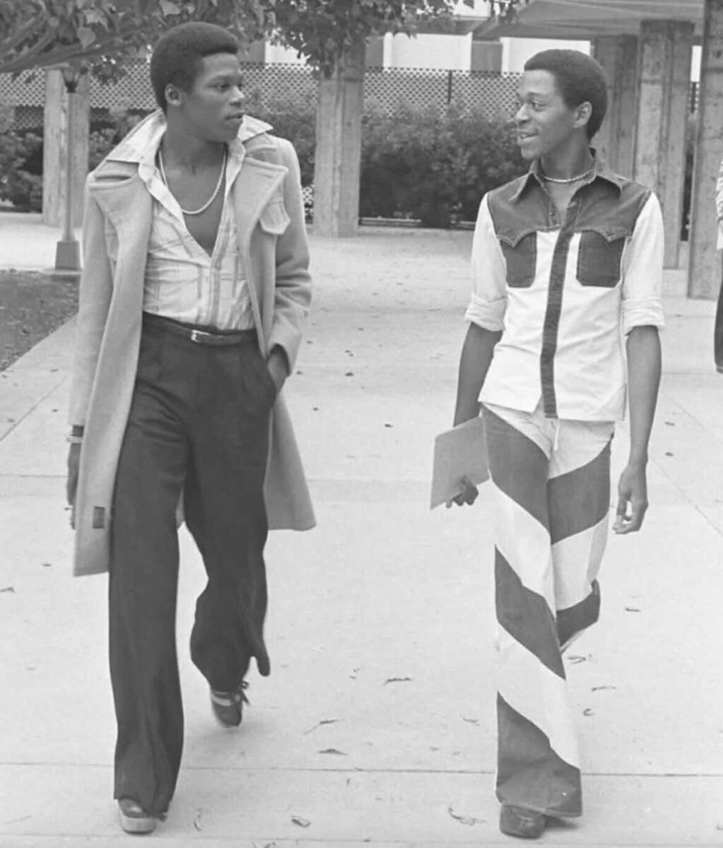 Two young men walk outside on a sidewalk. One wears a long coat, unbuttoned shirt, and flared pants. The other wears a matching shirt and wide-striped pants, holding a folder. Both are looking at each other and talking.