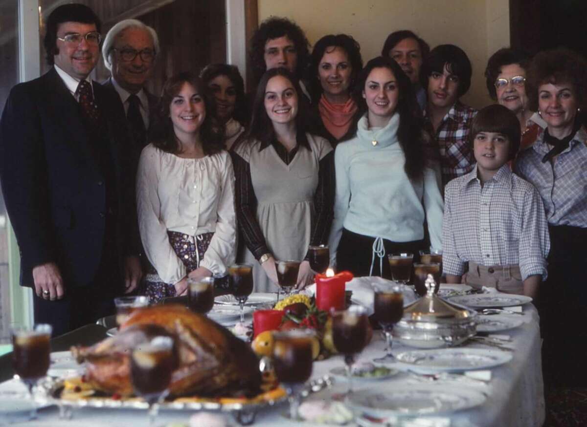 A group of people stand smiling behind a dinner table set with glasses, plates, candles, and a roast turkey, suggesting a festive family gathering or holiday meal.