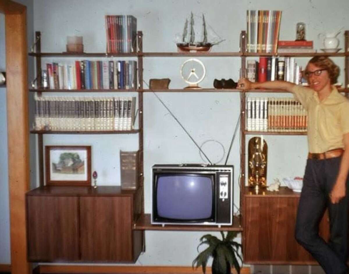 A person in glasses and a yellow shirt stands smiling next to a vintage wooden shelving unit with books, decor items, and a boxy TV. A model ship, clock, and plant are also visible on the shelves.