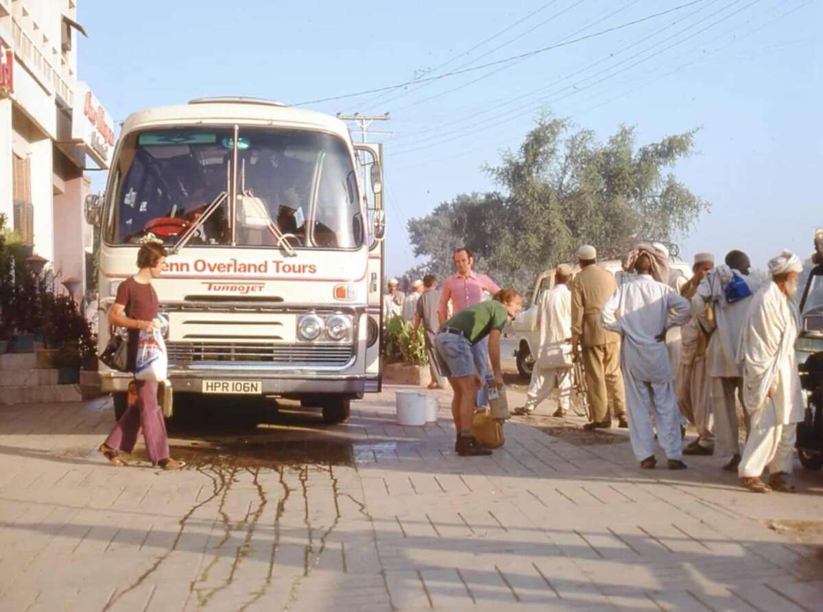 A group of people stands near a white tour bus labeled "Penn Overland Tours" on a sunny day. Some men wear traditional attire, while others wear casual Western clothing. Water is spilled on the pavement in front of the bus.