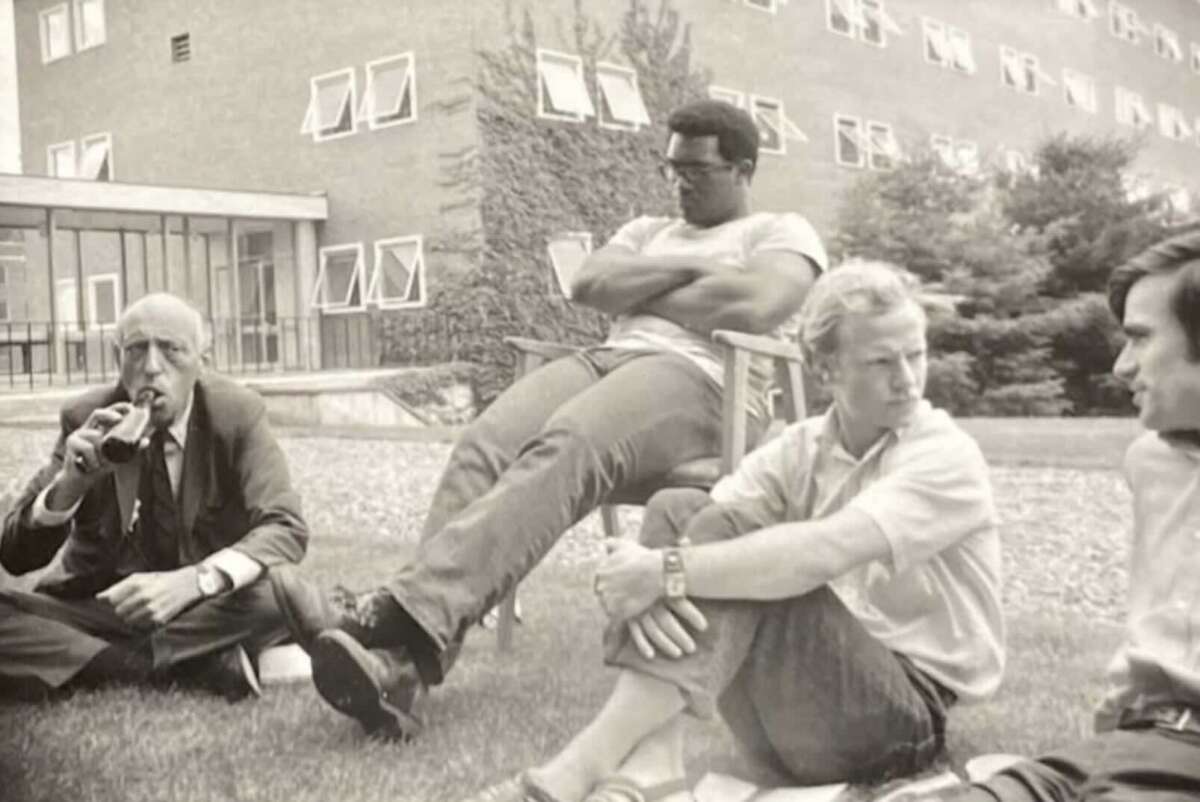 Four men sit outside near a building, three on the grass and one on a chair. One man in a suit drinks from a bottle, while the others wear casual clothes and appear to be engaged in conversation. Trees and windows are in the background.