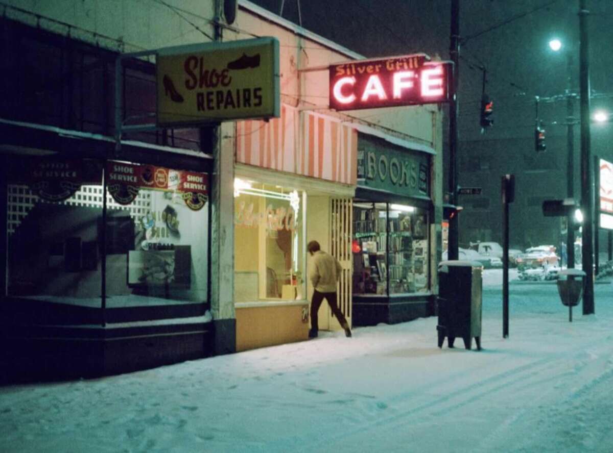 A person enters a brightly lit café at night on a snowy street, with neon signs for "Silver Grill Cafe" and "Shoe Repairs" glowing above. Snow covers the ground, and storefronts line the quiet street.