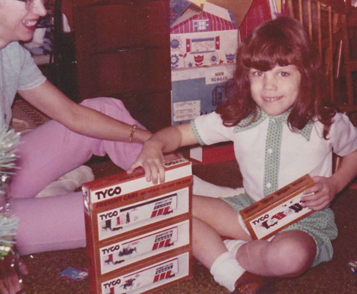 A young girl smiles while sitting on the floor, holding Tyco toy train boxes. An adult sits nearby, and wrapped presents are visible in the background, suggesting a festive occasion like Christmas.