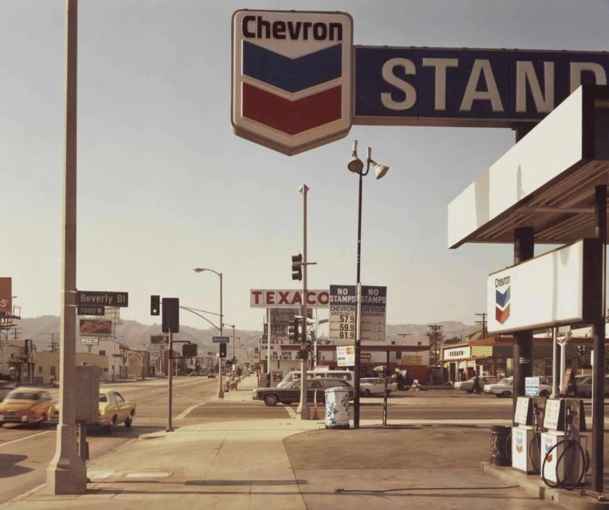 A vintage Chevron gas station on a sunny street corner with cars passing by. The price board and Texaco sign are visible, along with street signs for Beverly Blvd and La Brea Ave. Hills are seen in the background.