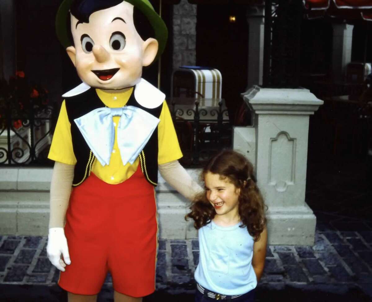 A young girl with curly hair smiles while standing next to a person in a Pinocchio costume on a paved area, with buildings and outdoor seating in the background.