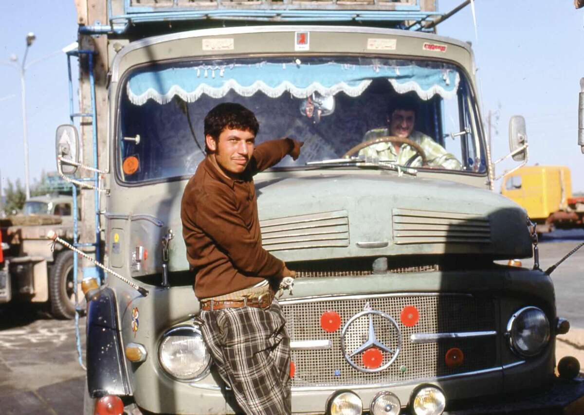 A man in plaid pants and a brown shirt leans against the front of a vintage Mercedes truck, smiling and pointing toward the driver inside, who is visible through the windshield. Other trucks are blurred in the background.