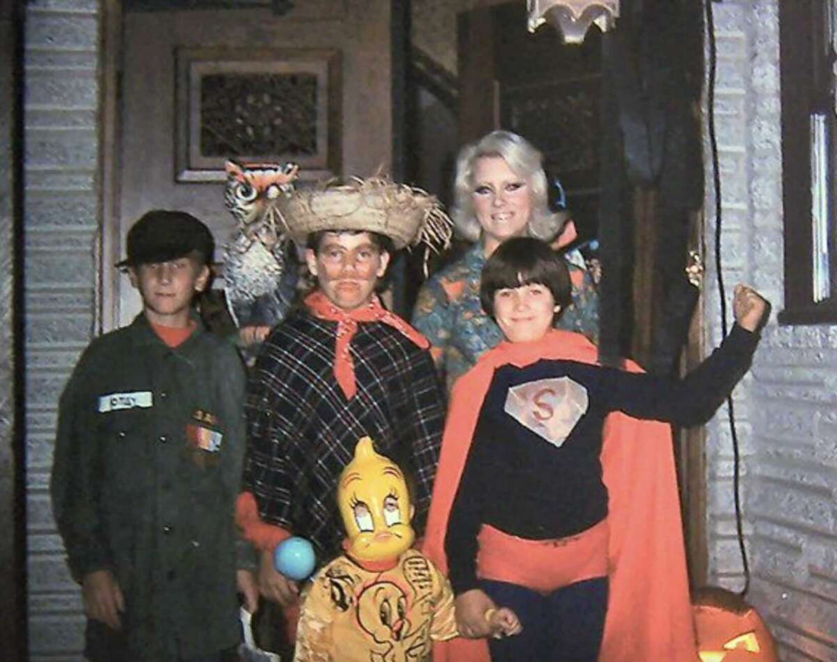 Four children in Halloween costumes and an adult stand on a porch. Costumes include a soldier, a scarecrow with a straw hat and plaid cape, a superhero with an "S" on the chest, and a Tweety Bird mask. A jack-o'-lantern is nearby.