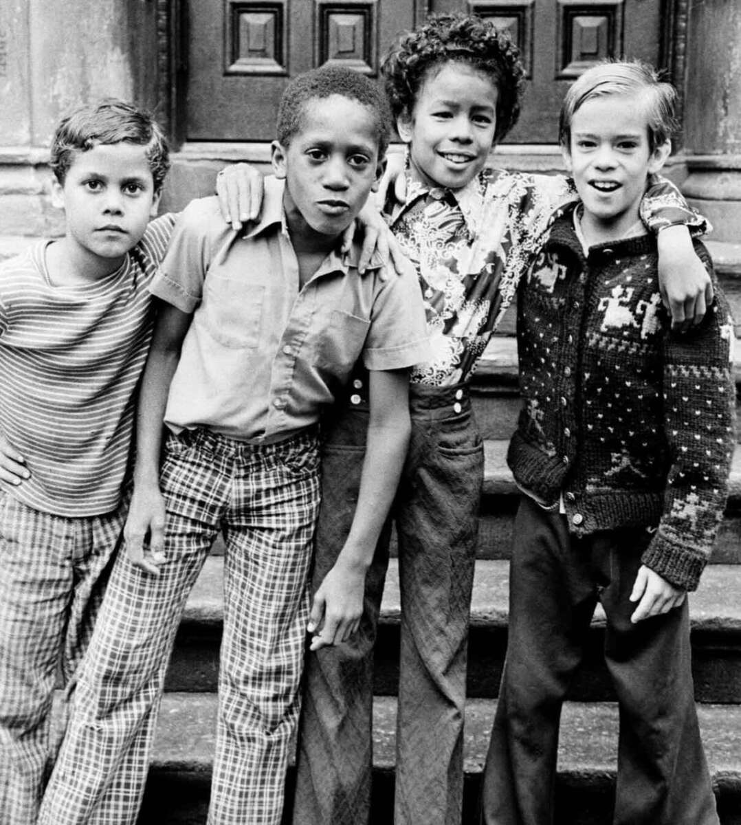 Four boys stand close together on stone steps, arms around each other’s shoulders, smiling at the camera. They wear patterned shirts and pants, reflecting a vintage or retro fashion style.