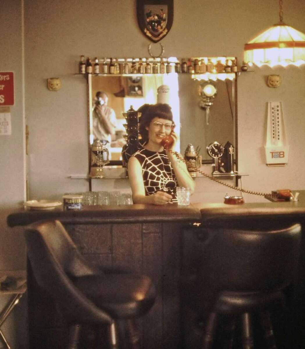 A woman in glasses and a patterned dress talks on a rotary phone behind a home bar. Various bottles and a mirror are behind her, with a man’s reflection visible. Two black bar stools are in the foreground.
