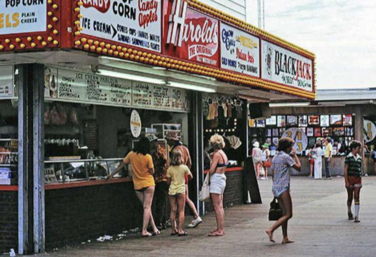 People stand in line at a boardwalk food stand under a marquee reading "Harold's" and "Black Jack," with signs for popcorn, ice cream, and candy apples. Others walk along the wooden boardwalk.