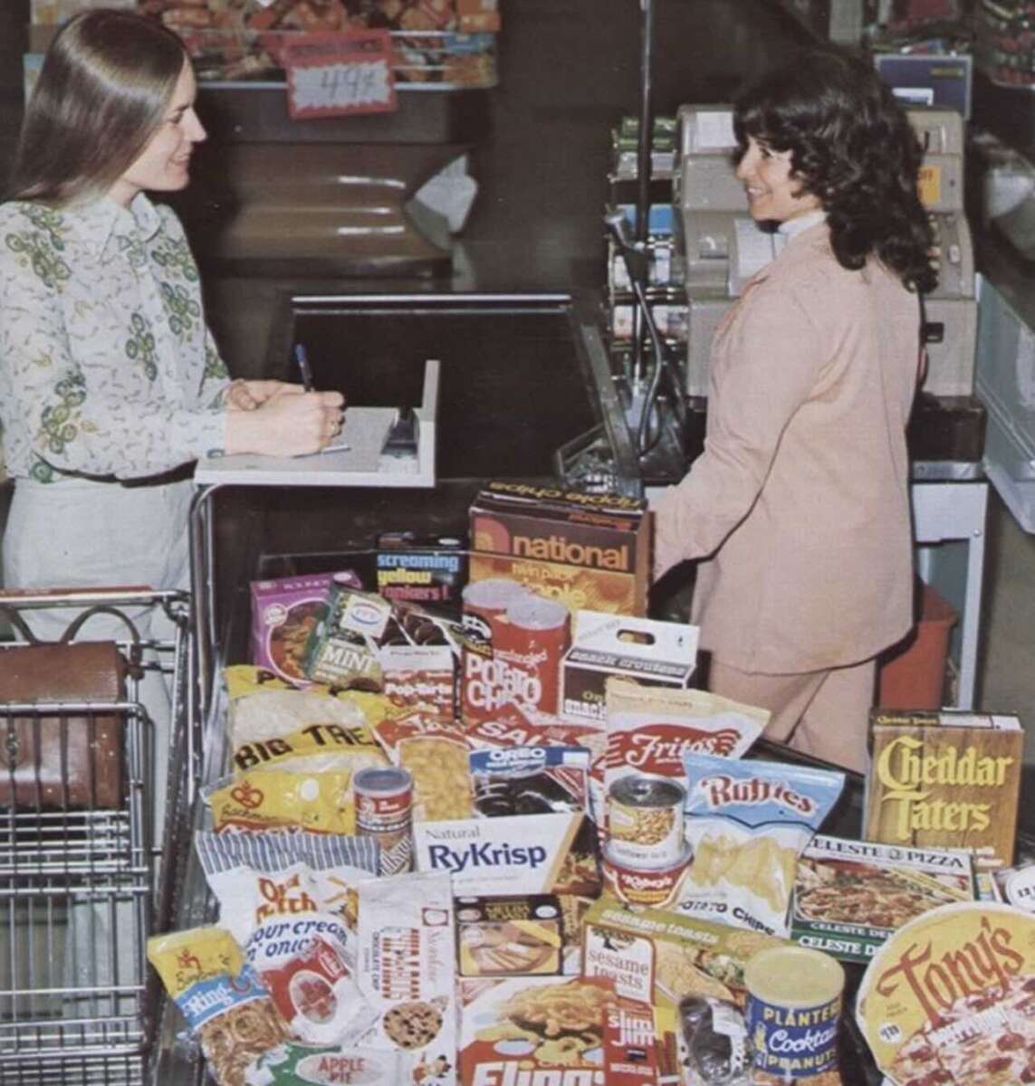 Two women stand at a grocery store checkout. One is writing on a notepad, the other stands at the register. Two shopping carts are filled with various food items and grocery products.