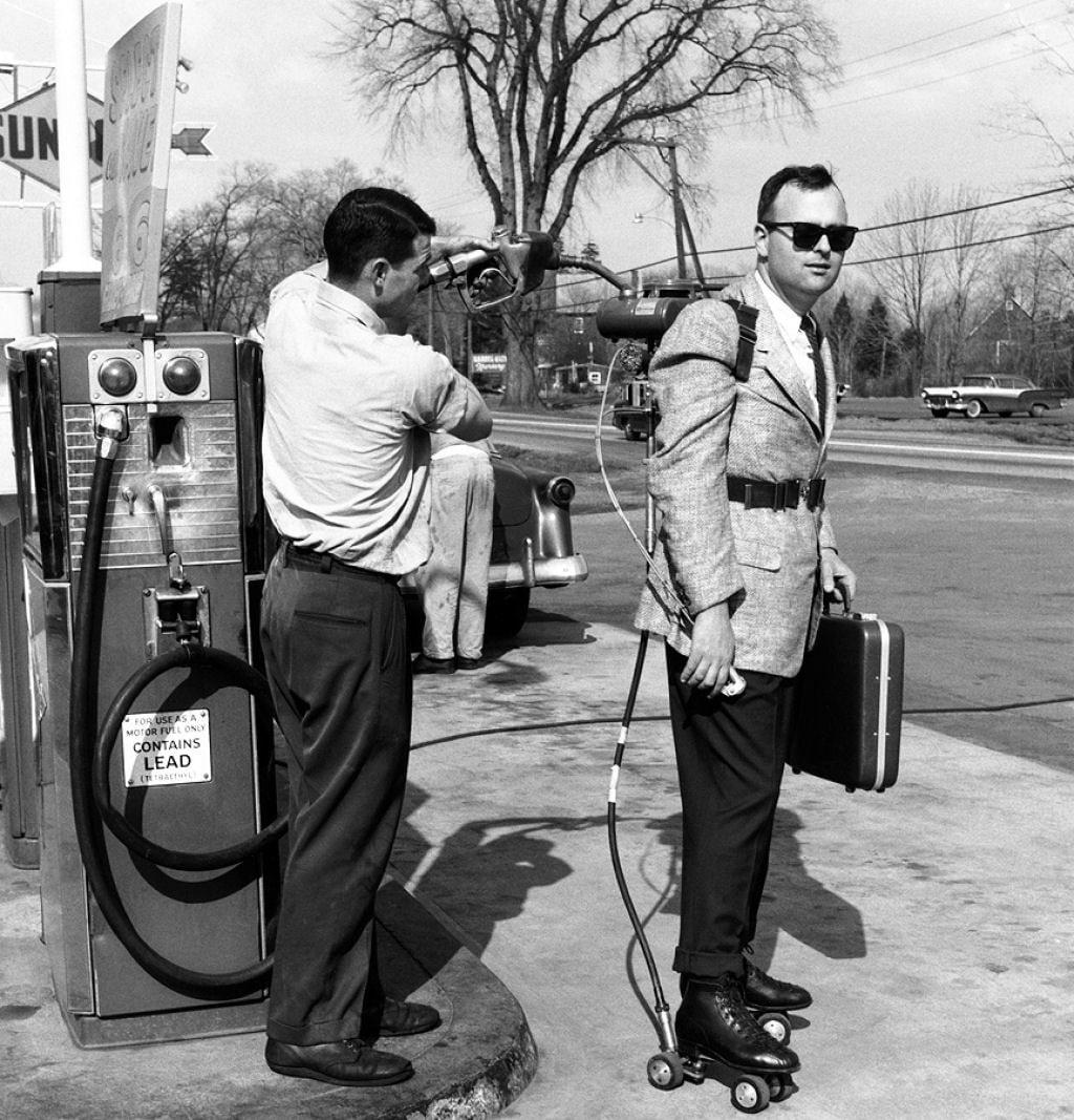 A man in a suit wearing roller skates and a jetpack stands at a gas station while another man fuels the jetpack. The man with the jetpack holds a briefcase, and trees and cars are visible in the background.