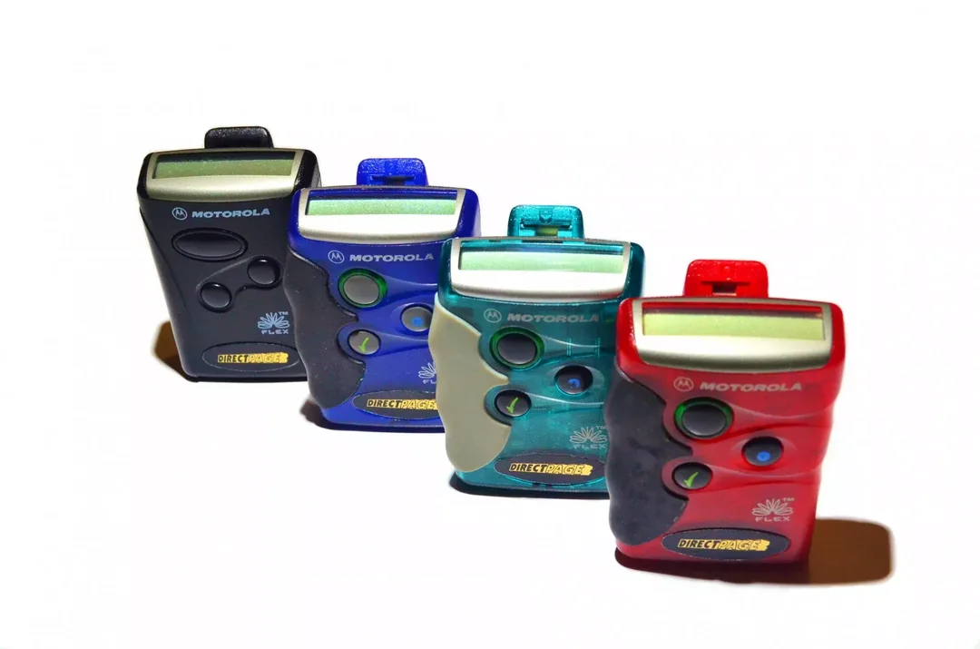 Four vintage Motorola pagers in black, blue, green, and red, are lined up diagonally on a white background, each with a small screen and buttons on the front.