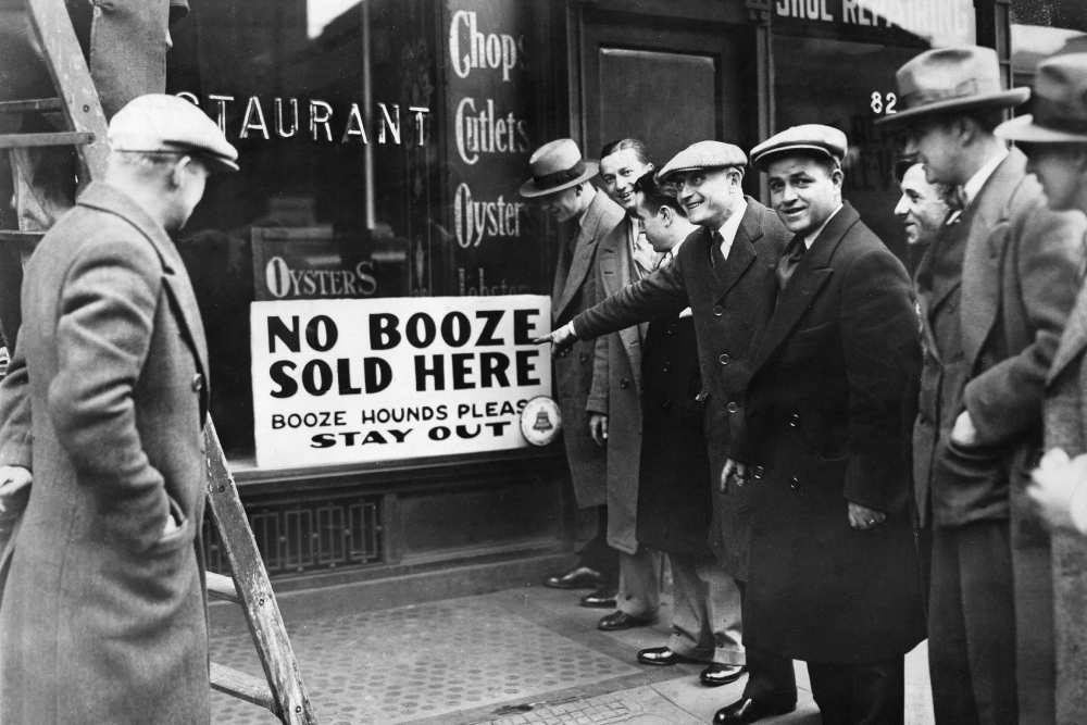 A group of men in coats and hats smile and gesture towards a restaurant window sign that reads, "NO BOOZE SOLD HERE. BOOZE HOUNDS PLEASE STAY OUT." The scene appears to be from the early 20th century.