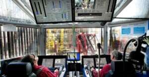 Two people sit at control panels inside a glass-enclosed room filled with monitors and screens, overlooking industrial machinery and equipment outside through large windows.