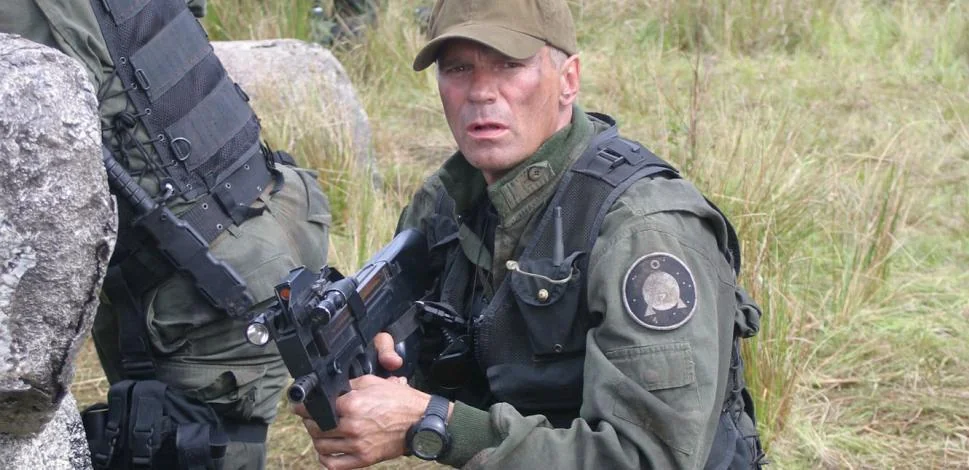 A man in tactical gear and a cap crouches outdoors, holding a rifle. He wears a dark vest with a circular patch on the arm, and is surrounded by grass and rocks.