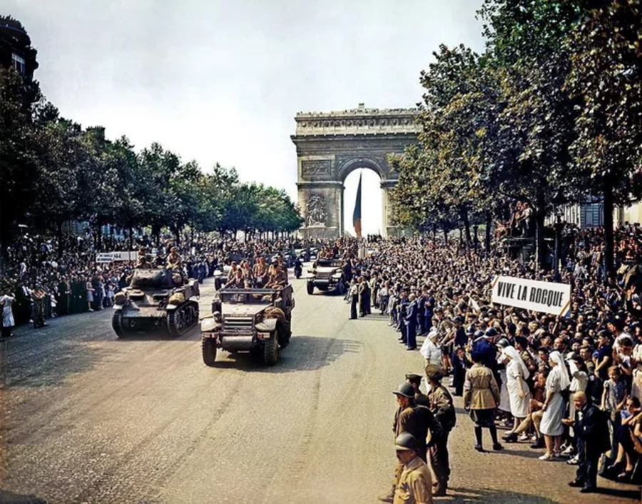 A historic military parade with tanks and vehicles moves down a wide street lined with crowds in front of the Arc de Triomphe. People are cheering and holding signs, including one that reads “Vive la Roque.”