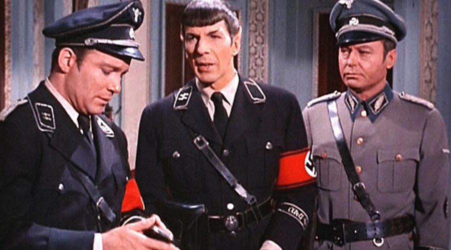 Three men dressed in Nazi-style uniforms stand indoors; one looks at an item in his hands, while the two others face forward. The scene appears to be from a film or television show.