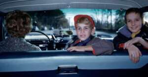 Two children sit in the back seat of a car, smiling at the camera, while an adult drives. The view is from the backseat, showing a road and trees through the windshield.