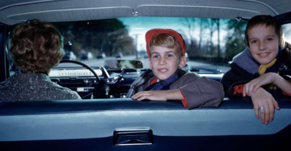 Two children sit in the back seat of a car, smiling at the camera, while an adult drives. The view is from the backseat, showing a road and trees through the windshield.